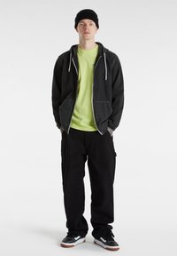 Musta huppari valkoisilla ompeleilla, neonvihreä t-paita, mustat cargo-housut ja mustat lenkkarit valkoisilla yksityiskohdilla. Yksinkertainen pipo päässä.