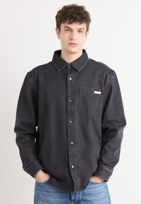 Black denim shirt with a button-down collar, chest pocket, and white button closures. Features a relaxed fit and subtle stitching details.