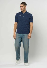 Férfi sötétkék galléros pólóban, világoskék farmerral és fehér sportcipőben, egyszínű világos háttér előtt áll, balra néz.