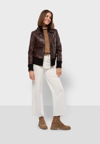 Braune Lederbomberjacke mit gerippten Bündchen, kombiniert mit einem hellbraunen Rollkragenpullover, weißen weit geschnittenen Hosen und braunen Schnürstiefeletten.