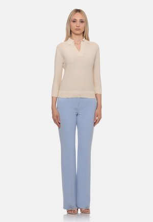 Donna bionda in piedi che indossa un top beige lavorato a maglia con colletto e pantaloni larghi azzurri su sfondo bianco.