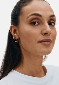 Frau mit zurückgebundenen dunklen Haaren trägt ein weißes Oberteil und drei verschiedene goldene Ohrringe am linken Ohr, darunter einen Spike, einen Edelstein und einen sternförmigen Stecker.