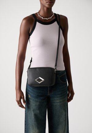 Vrouw in een lichtpaarse tanktop met zwarte accenten, wijdvallende vervaagde spijkerbroek, een zilveren ketting en een zwarte schoudertas met een zilveren logo.