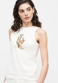 Γυναίκα με καστανά σγουρά μαλλιά που φοράει λευκό αμάνικο μπλουζάκι με γραφικό σχέδιο κάκτου και μποτάκι καουμπόη, στέκεται μπροστά σε απλό φόντο.