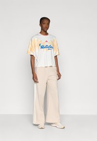 Fehér rövid ujjú póló narancssárga geometriai mintával és kék "Adidas" felirattal, világos bézs bő szárú nadrággal és fehér tornacipővel párosítva.