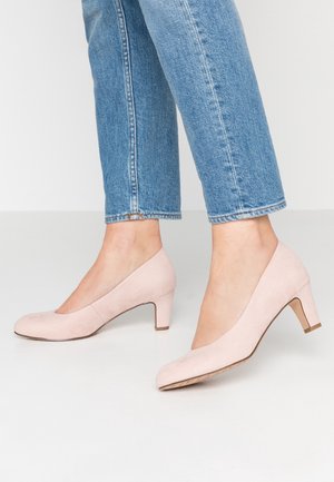 Des escarpins en suède rose clair avec un bout rond et un petit talon carré. La chaussure a une texture lisse et un design légèrement incliné.