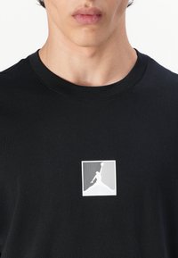 Melnas kokvilnas T-kreklis ar balto Air Jordan logo kvadrātā uz krūšu. Vienkāršs apaļais kakla dizains ar īsām piedurknēm.