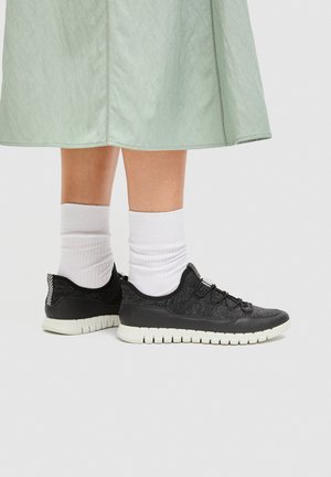 Legs wearing white socks and black athletic shoes with white soles, standing against a plain background beneath a green skirt.