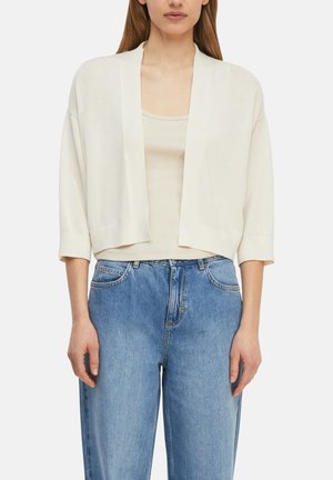 Frau trägt eine cremefarbene, offene Strickjacke über einem beigen Tanktop, kombiniert mit hoch taillierten hellblauen Jeans.