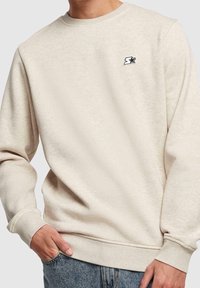 Homme portant un sweat-shirt beige à manches longues avec un logo étoile noir et blanc sur la poitrine, associé à un jean en denim bleu.