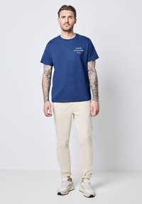 Náměšťová modrá bavlněná tričko s krátkým rukávem, s bílým potiskem. V kombinaci s světle béžovými kalhotami a šedými teniskami.