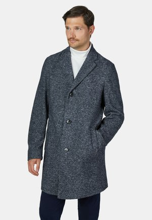 Man draagt een midlange donkergrijze wollen jas met knopen en zakken, een witte coltrui en een donkere broek, kijkt iets opzij.