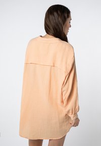 Pfirsichfarbenes oversize Hemd aus strukturiertem Stoff, mit einem fließenden Design, langen Ärmeln und einer Rückentasche.
