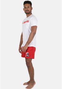 Hvid t-shirt med røde vandrette striber, parret med røde shorts med et palmetræ-grafik og logo. Model står barfodet.
