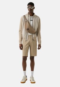 Veste bomber beige clair, t-shirt blanc avec logo, shorts kaki et baskets blanches avec semelles beige. Lunettes de soleil ovales et sac en bandoulière beige.