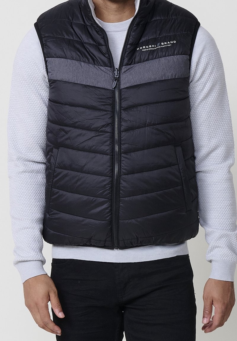 Homme portant une veste matelassée noire et grise par-dessus un pull gris clair texturé à manches longues et un pantalon noir, les bras détendus le long du corps.