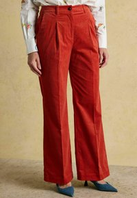 Sarkani kordbārdu bikses ar platu kāju un dubultām krokām priekšpusē. Stylistētas kopā ar zilām smailām kurpēm uz papēžiem. Smalki ribota tekstūra.