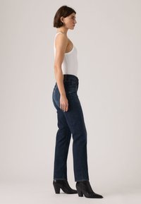 Γυναίκα με λευκό αμάνικο τοπ, σκούρο μπλε τζιν ψηλόμεσο και μαύρα μποτάκια με τακούνι, όρθια πλάγια σε απλό φόντο.