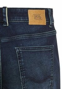 Jean en denim bleu marine avec une finition texturée, portant une étiquette en cuir beige avec l'inscription "camel active" et des couture jaunes contrastantes.