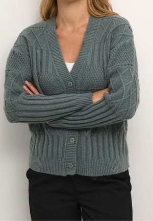 Cardigan en tricot vert avec un col en V, motifs texturés sur les manches, ourlet côtelé et boutons sur le devant. Stylé avec un pantalon noir.