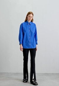 Μπλε πουκαμίσα με μακριά μανίκια, φορεμένη με μαύρο παντελόνι με πλαϊνές σχισμές και μαύρες πλατφόρμες. Απλός σχεδιασμός, ομαλή υφή.