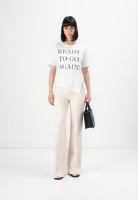 Γυναίκα με ίσια μαύρα μαλλιά, φορώντας t-shirt λευκού χρώματος που γράφει "ΕΤΟΙΜΗ ΓΙΑ ΑΛΛΗ ΜΙΑ ΦΟΡΑ!", μπεζ παντελόνι, μαύρες σαγιονάρες και κρατώντας μαύρη τσάντα, όρθια σε άσπρο φόντο.