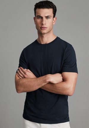 Unge mand med mørkt krøllet hår iført en sort kortærmet skjorte og hvide bukser, stående med armene over kors mod en grå baggrund.