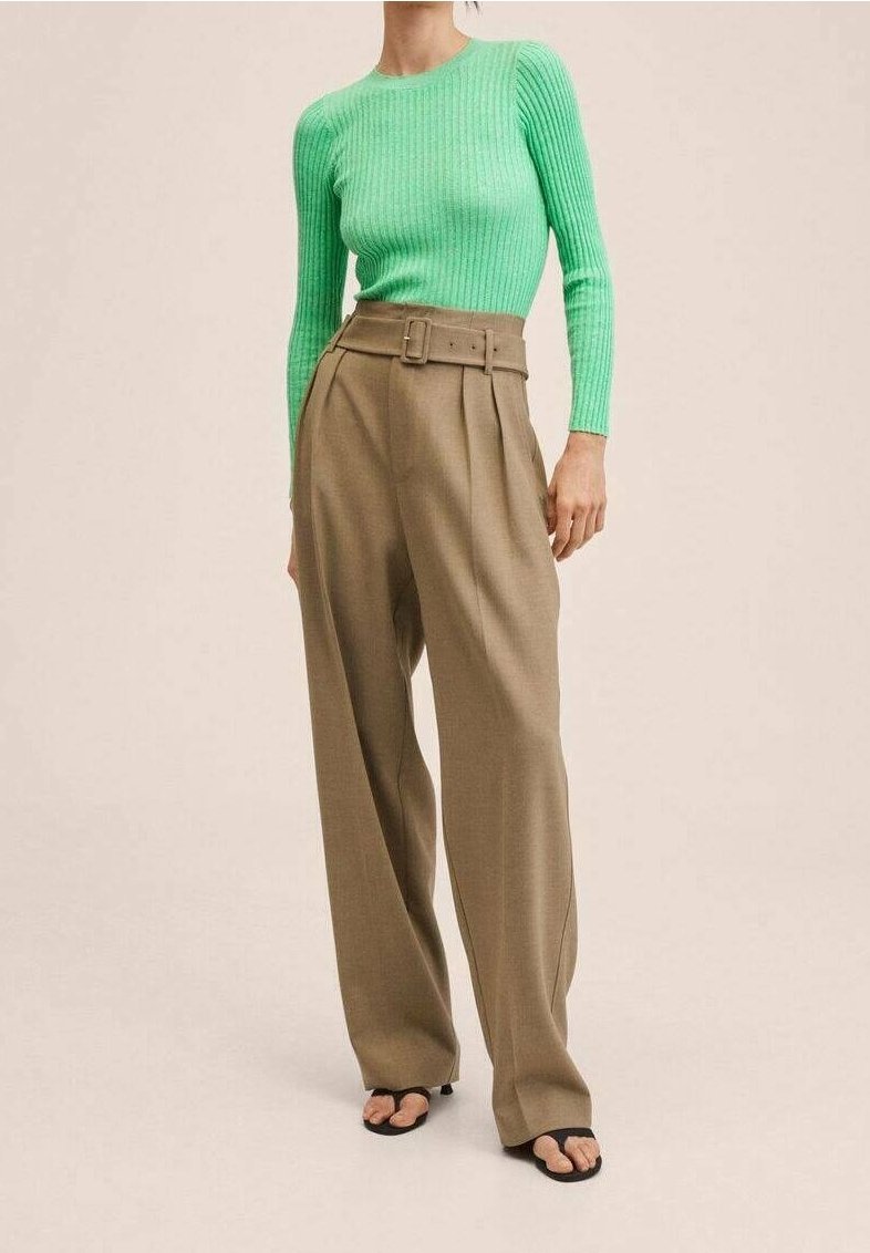 Grün gerippter Pulli, hochtaillierte beigefarbene plissierte Hose mit Gürtel und schwarze Flip-Flops vor einem neutralen Hintergrund.