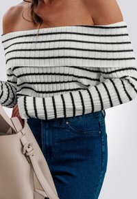 Maglione a spalle scoperte a righe nere su sfondo bianco. Presenta una texture a costine, abbinato a denim blu e una borsa beige chiaro.