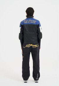 Läderjacka i svart och blått med vita detaljer, som har en broderad "Ed Hardy"-logotyp på ryggen och skyddande vaddering på armarna.