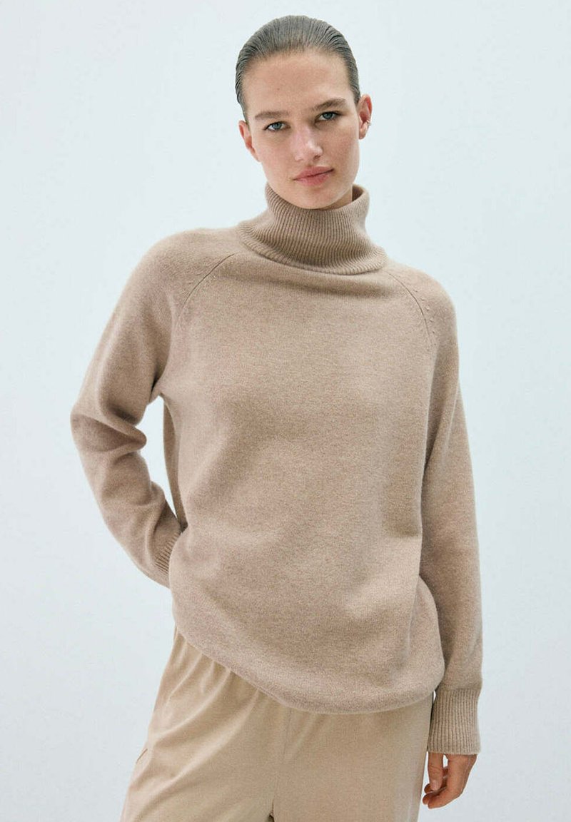 Femme aux cheveux tirés en arrière, portant un pull à col roulé beige et un pantalon beige clair assorti sur un fond clair uni.
