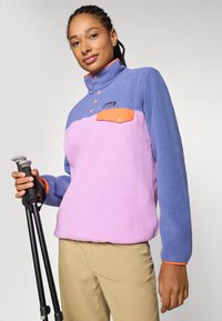 Fleece pullover in purple and pink with an orange collar; features a chest pocket and contrasting hardware. Model holds trekking poles.