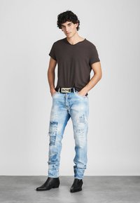Lichtblauwe distressed jeans met gescheurde details, gecombineerd met een donkerbruin t-shirt en een zwarte riem met een gouden gesp. Zwarte enkellaarsjes.
