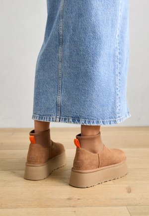 Personne portant une jupe en jean bleu clair et des bottines compensées beiges avec des languettes orange, debout sur un sol en bois.