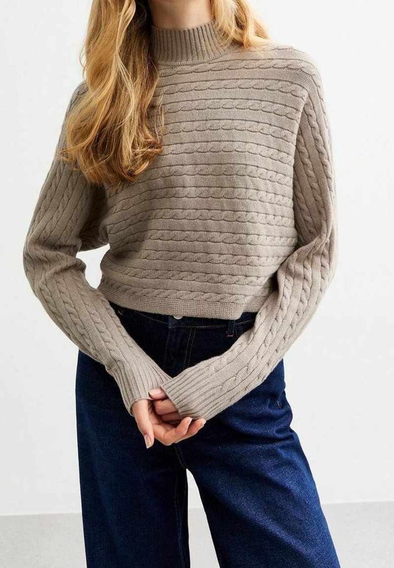 Cremefarbenes Strickpullover mit strukturierten Zopfmustern, hohem Kragen und langen Ärmeln. Kombiniert mit dunkelblauen, weit geschnittenen Jeans.