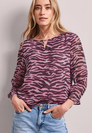 Femme blonde portant une blouse rose et violet foncé à rayures tigre avec des manches ballon transparentes et un jean bleu clair, les mains dans les poches.