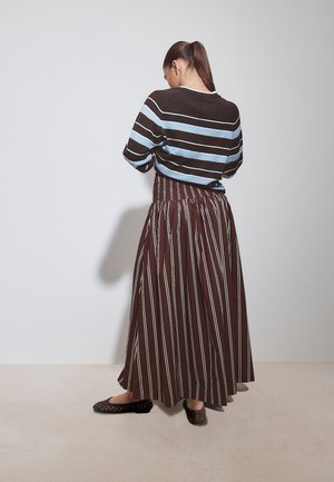 Woman with brown ponytail wearing a brown and light blue striped sweater and a long brown skirt with vertical white stripes, standing against a white wall.