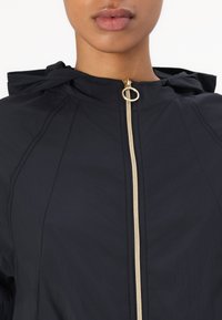 Giacca a vento nera con cappuccio e zip dorata, realizzata in tessuto con texture e dettagli delle cuciture, progettata per offrire comfort e stile leggero.