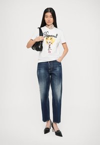 T-shirt grafica bianca con illustrazione, jeans blu a gamba dritta e scarpe con tacco nere. La modella tiene una borsa nera.