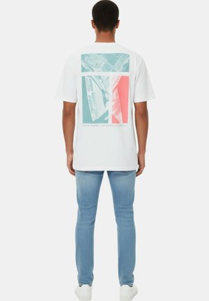 Homme portant une chemise blanche à manches courtes avec un motif abstrait turquoise et corail dans le dos, associée à un jean bleu clair et des baskets blanches, debout.
