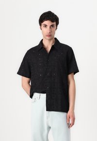 Chemise noire à manches courtes avec un design texturé brodé incluant des motifs découpés et un devant à boutons. Associée à un jean en denim clair.