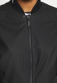 Černá zipová bunda s ribovým límcem a viditelným jezdcem, nošená přes pruhované tričko, střih krátký na krku a ramenou.