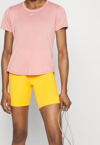 Top deportivo rosa de manga corta con un logo blanco, combinado con pantalones cortos ajustados amarillos brillantes. Sosteniendo una cuerda para saltar. Textura suave.