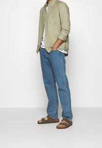 Lichtgroen overhemd met knopen, witte T-shirt, blauwe rechte jeans en bruine sandalen met twee banden en kurken zolen.