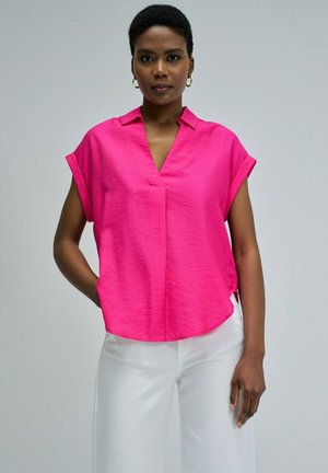 Mujer que lleva una blusa rosa brillante de manga corta con escote en V y pantalones blancos, de pie con una mano en el bolsillo contra un fondo liso.