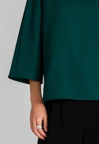Top verde, comodo e largo con maniche a tre quarti, abbinato a pantaloni neri. La mano presenta un anello d'argento con un design intrecciato.