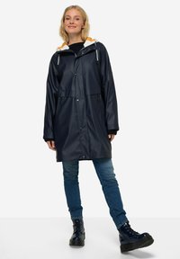 Voděodolný námořnický pláštěnka s bílou a oranžovou kapucí, patentkami, postranními kapsami a dlouhými rukávy. Nosit s modrými džíny a černými botami.