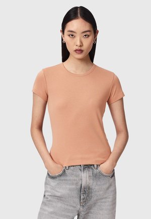 Kvinde med glat sort hår iført ferskenfarvet t-shirt med korte ærmer og lysegrå højttaljede jeans står med hænderne i lommerne mod en grå baggrund.