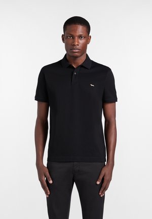 Uomo che indossa una polo nera a maniche corte con un piccolo logo di un cane sul petto a sinistra, abbinata a pantaloni neri, in piedi davanti a uno sfondo bianco.