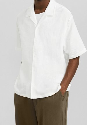 Homme portant une chemise blanche à manches courtes texturée boutonnée par-dessus un sous-vêtement blanc avec un pantalon vert olive, une main dans la poche.